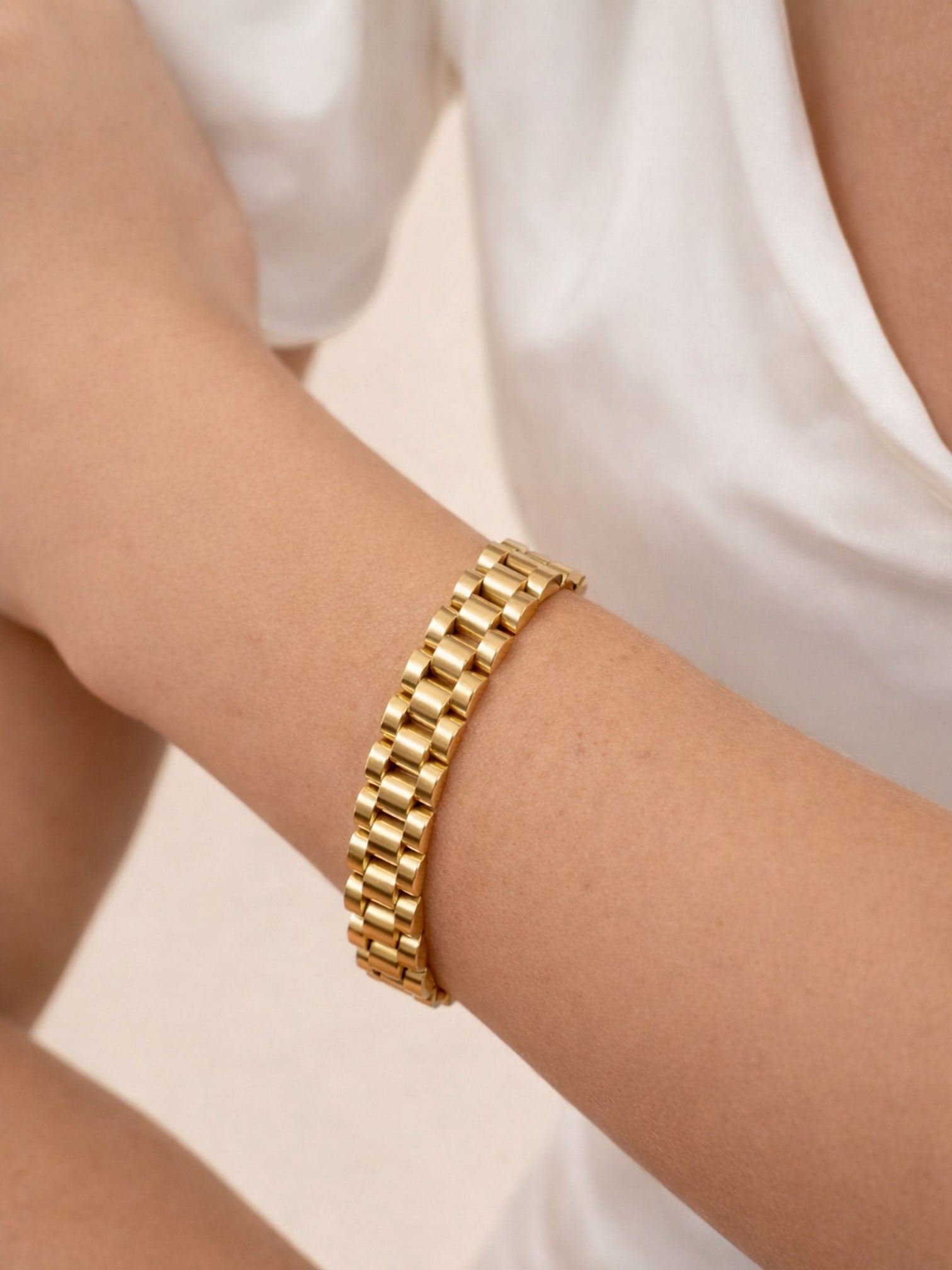 Gold watchband-style bracelet on wrist, featuring polished rectangular links in a rich 18K gold tone, worn against a soft neutral background.