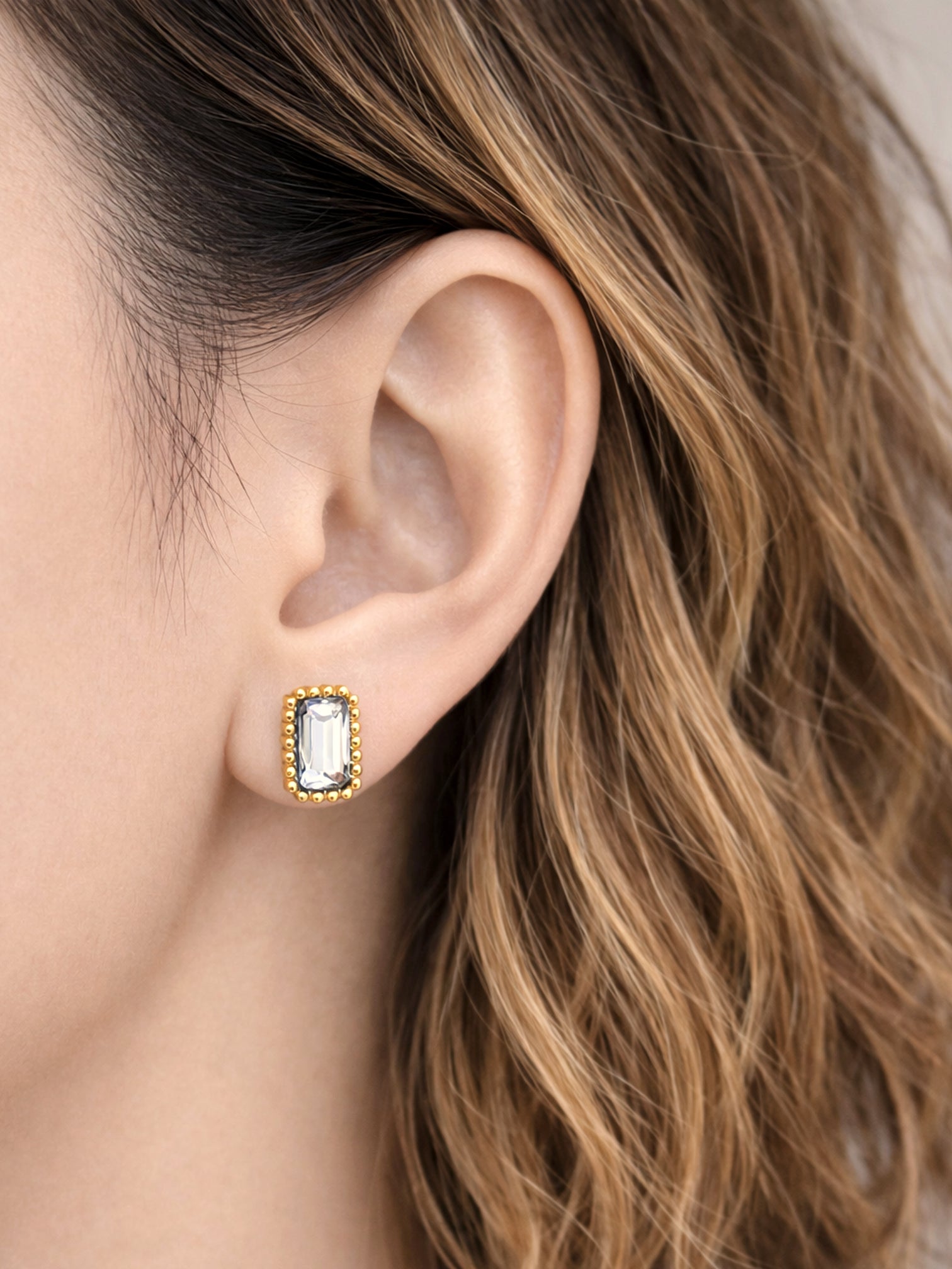 Aurae screwback earrings featuring a rectangular clear baguette-cut stone set in a gold beaded frame, worn on the ear in a close-up lifestyle image.