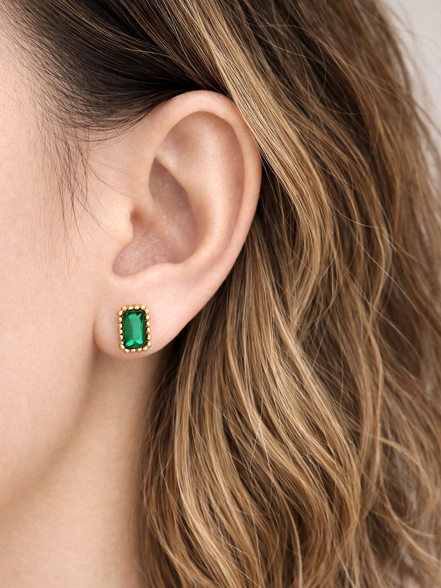 Aurae screwback earrings featuring a rectangular emerald-green gemstone set in a gold beaded frame, worn on the ear in a close-up lifestyle image.