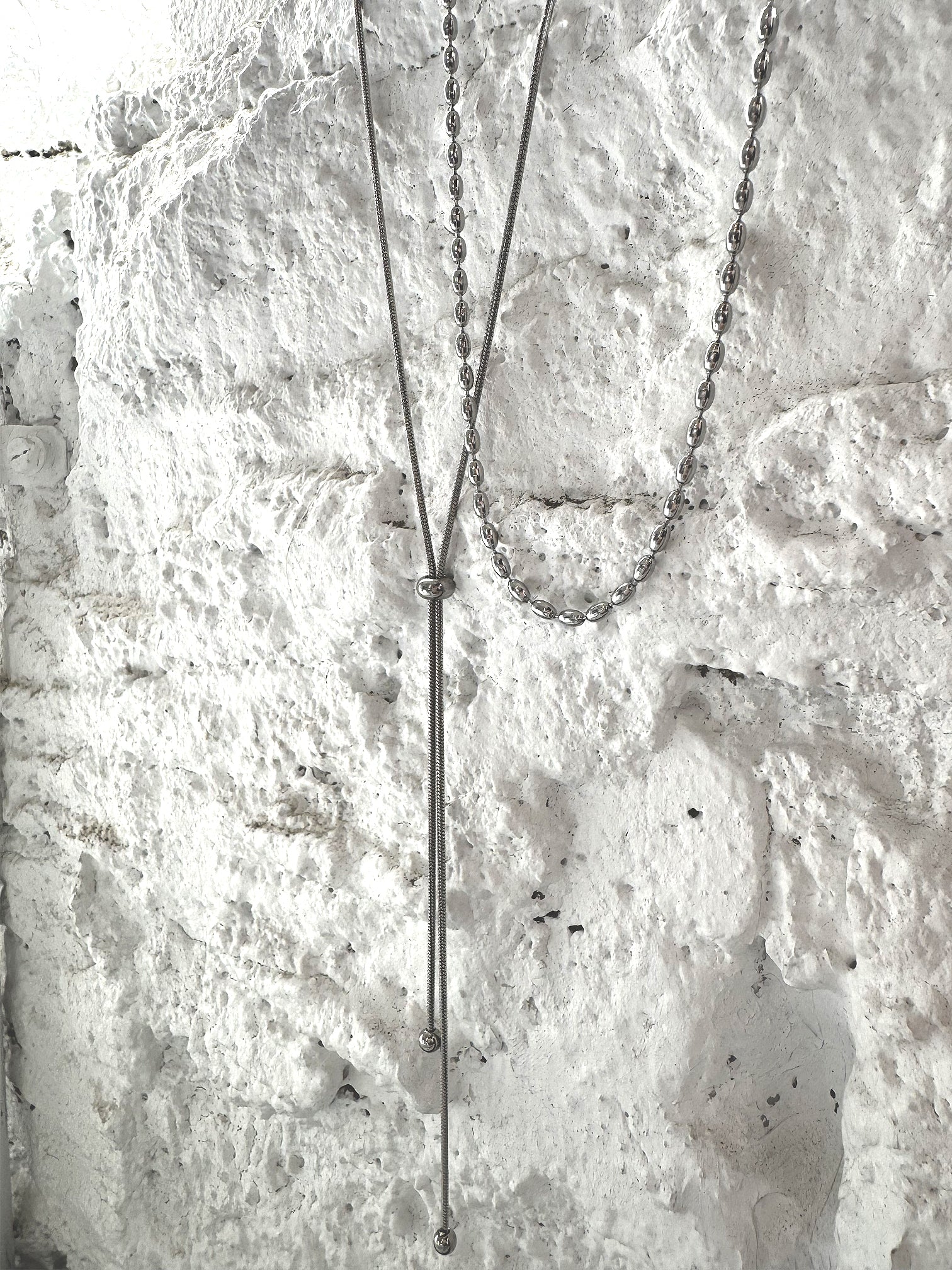 Silver adjustable Y lariat necklace with dual chain layers, featuring a sleek snake chain drop and polished oval link chain, set against textured white stone background.