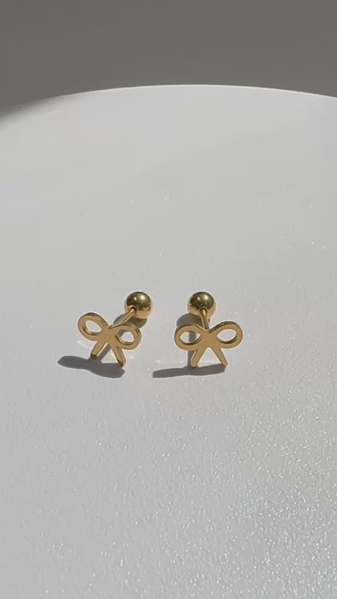 Minimal bow-shaped Bree stud earrings rotating on a turntable, crafted with 18K gold plating over titanium steel, showcasing a delicate open bow design with a polished, lightweight finish on a clean white surface. | myEdzy